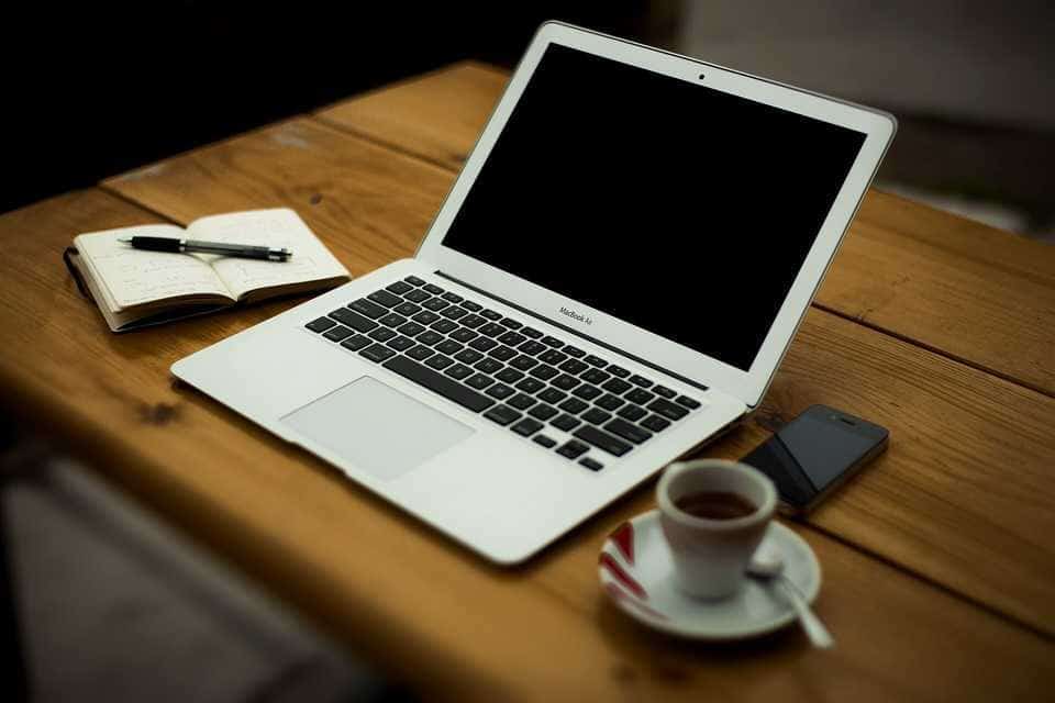 A laptop, notebook, phone and coffee on a wooden desk